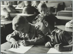 Kinderen met pen en papier in de schoolbank in de klas. [1937]. Plaats onbekend.